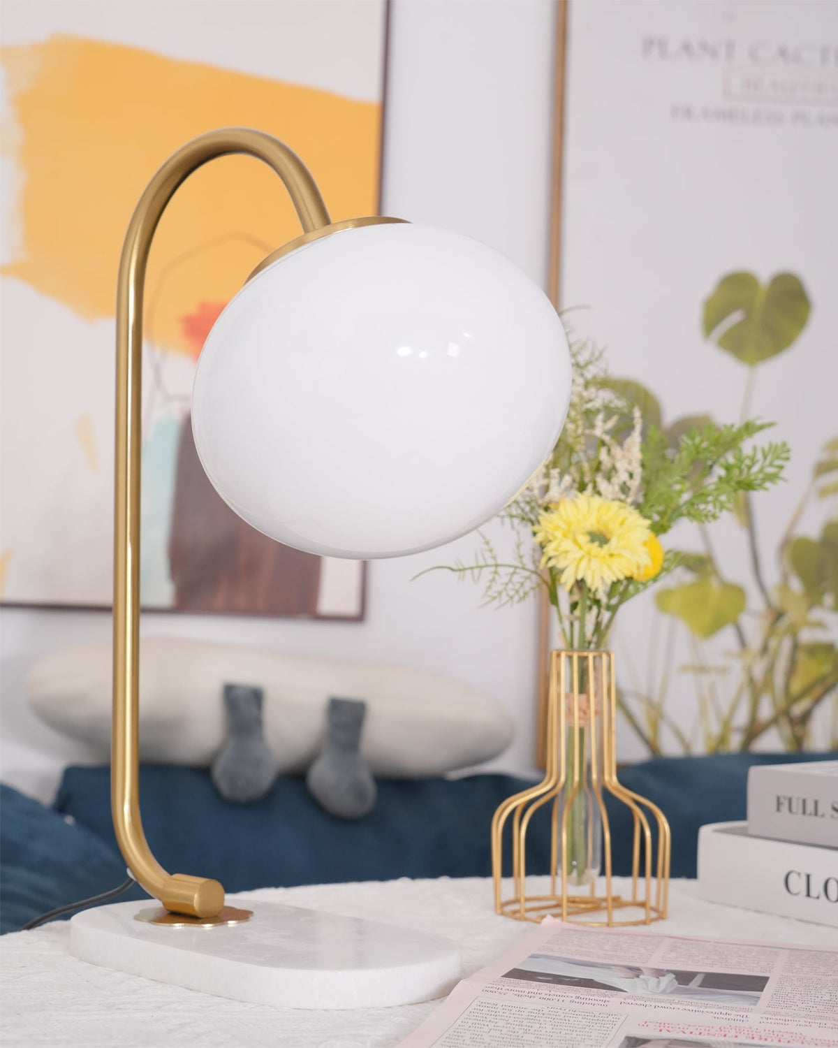 A lit Clérachic table lamp with gold arm and white shade stands against sheer curtains, wire baskets of flowers on the table beside it.