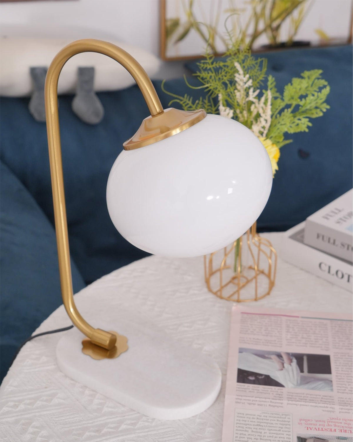 Close view of the Clérachic table lamp's gold arm and white shade beside a blue cushioned sofa arm, greenery softly out of focus.