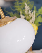 Extreme close-up of the Clérachic table lamp's glossy white glass shade and gold cap, sprigs of greenery visible just behind it.