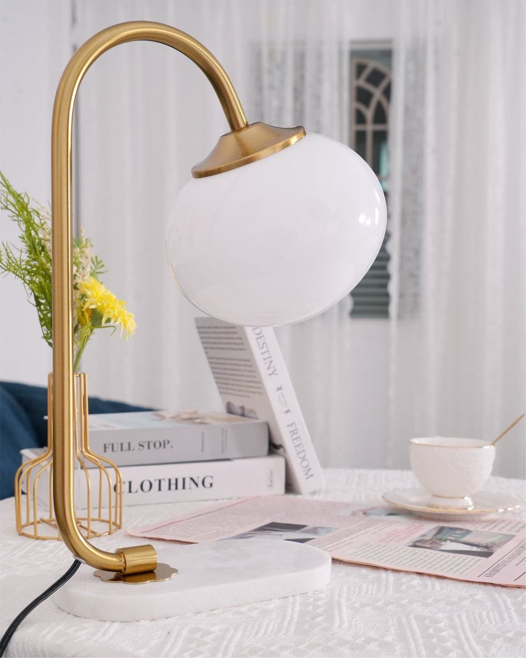 The Clérachic table lamp's white shade glows on a gold arm beside a wire basket of daisies and a stacked magazine on a marble base.