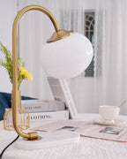 The Clérachic table lamp's white shade glows on a gold arm beside a wire basket of daisies and a stacked magazine on a marble base.