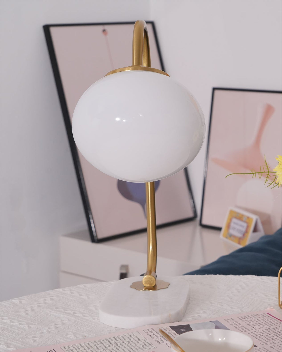 An unlit Clérachic table lamp with gold arm and white shade stands before framed art and sheer curtains on a marble-topped table.