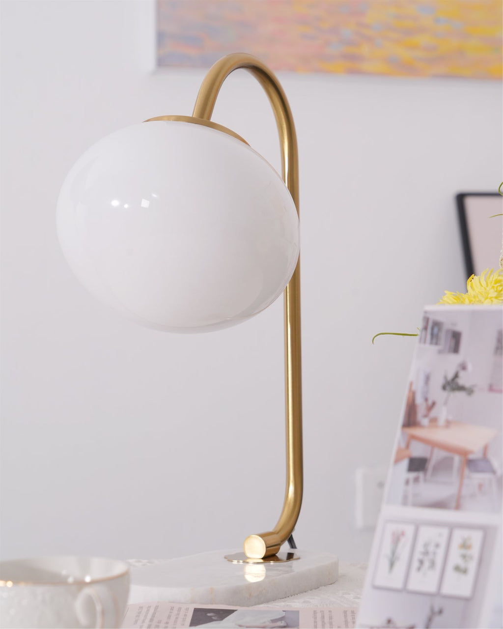 The Clérachic table lamp's white shade sits unlit on a gold arm above a marble base, a teacup and stacked magazines nearby on the table.