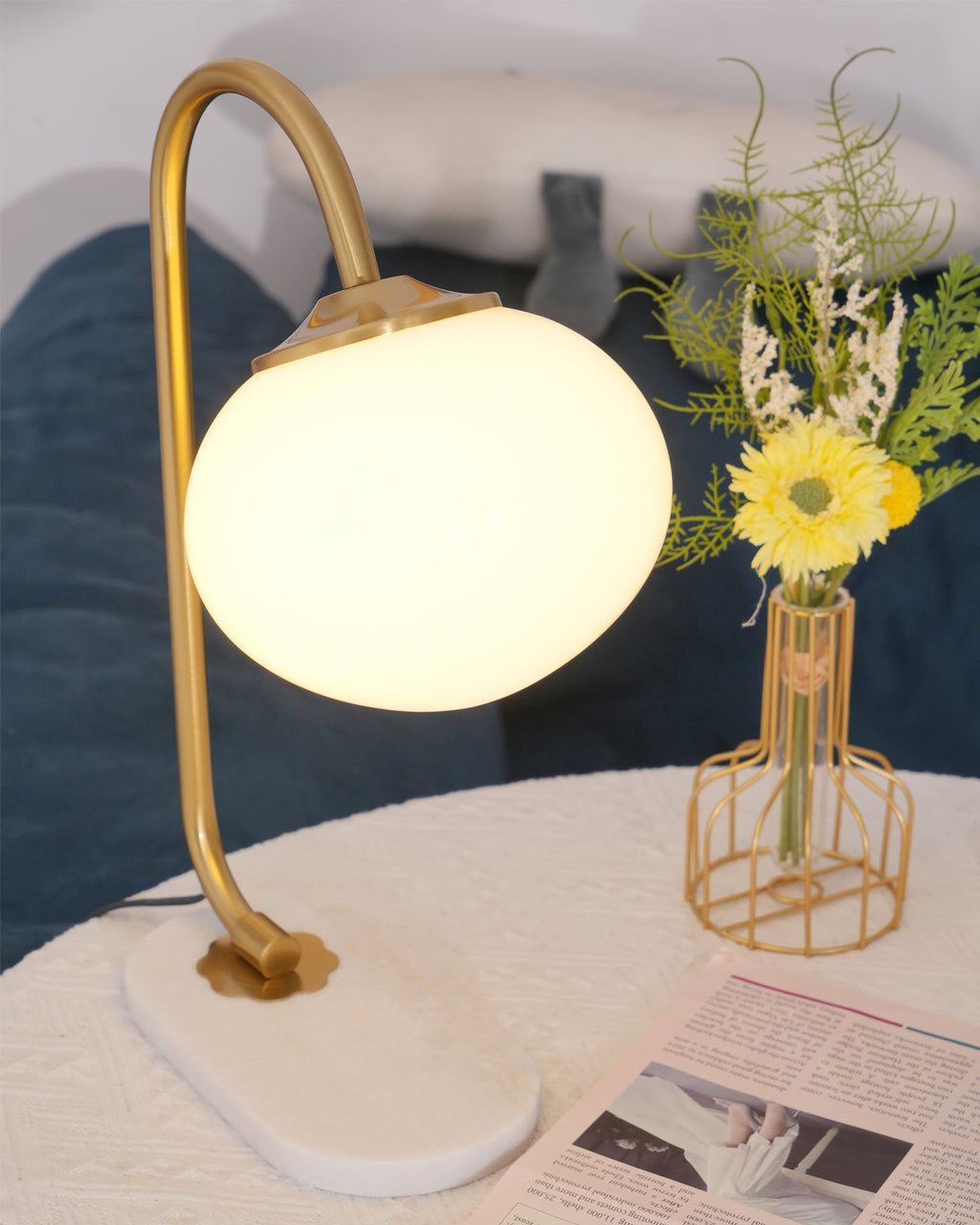 A lit Clérachic table lamp with gold arm and white shade stands on marble beside a blue sofa cushion and a wire basket of daisies.