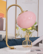 The Clérachic table lamp's pink shade sits unlit on a gold arm and marble base, beside a wire basket of daisies on a blue cushion.