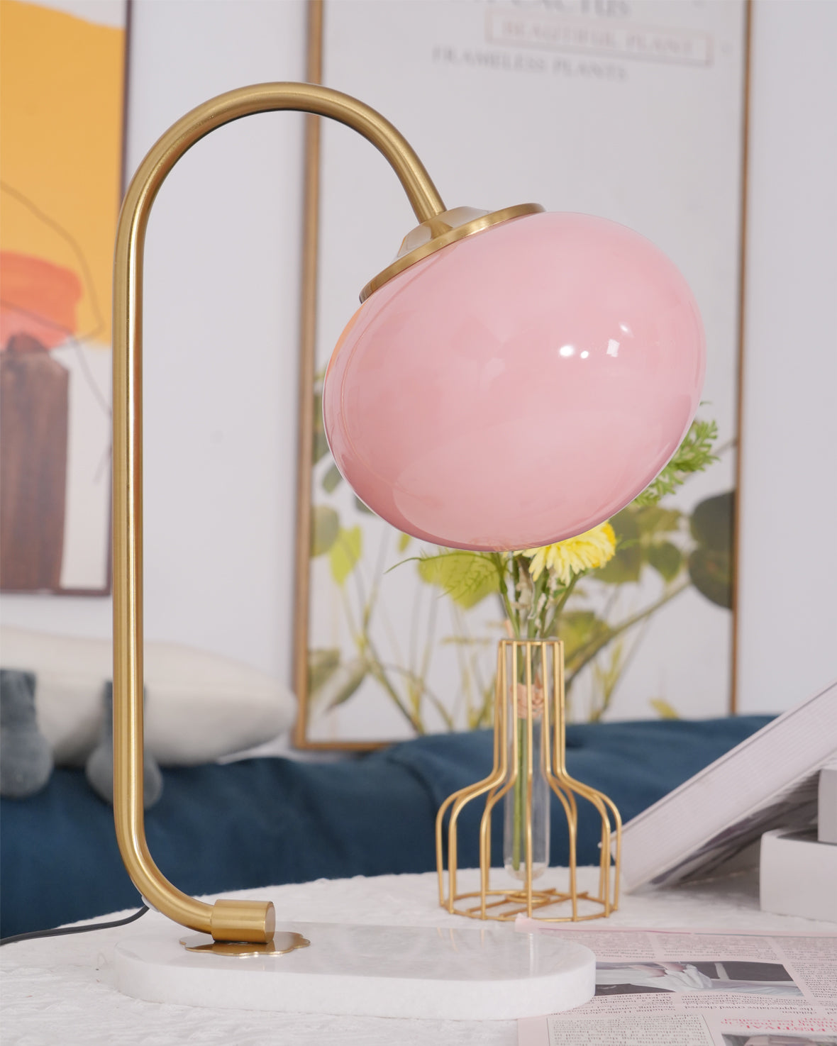 The Clérachic table lamp's pink shade sits unlit on a gold arm and marble base, beside a wire basket of daisies on a blue cushion.