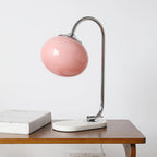 A pink-shaded Clérachic table lamp on a chrome arm rests unlit at the edge of a wooden table, stacked books positioned beneath it.