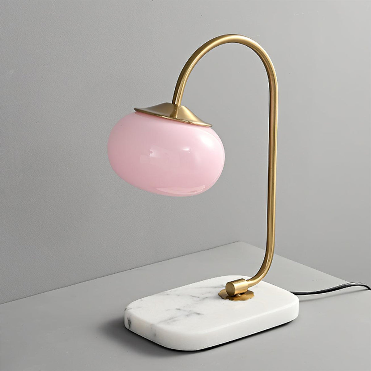 Clérachic pastel glass table lamp with a pink globe shade on a gold arm, standing on a rectangular white marble base against grey.