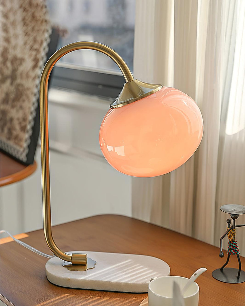 The Clérachic table lamp's pink shade sits on a sunlit wooden table, brass arm beside a small pitcher and a wire figurine ornament.