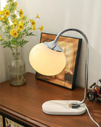 An off-white glass shade lights the Clérachic table lamp's chrome arm, set on a wooden dresser beside a framed picture and yellow daisies.