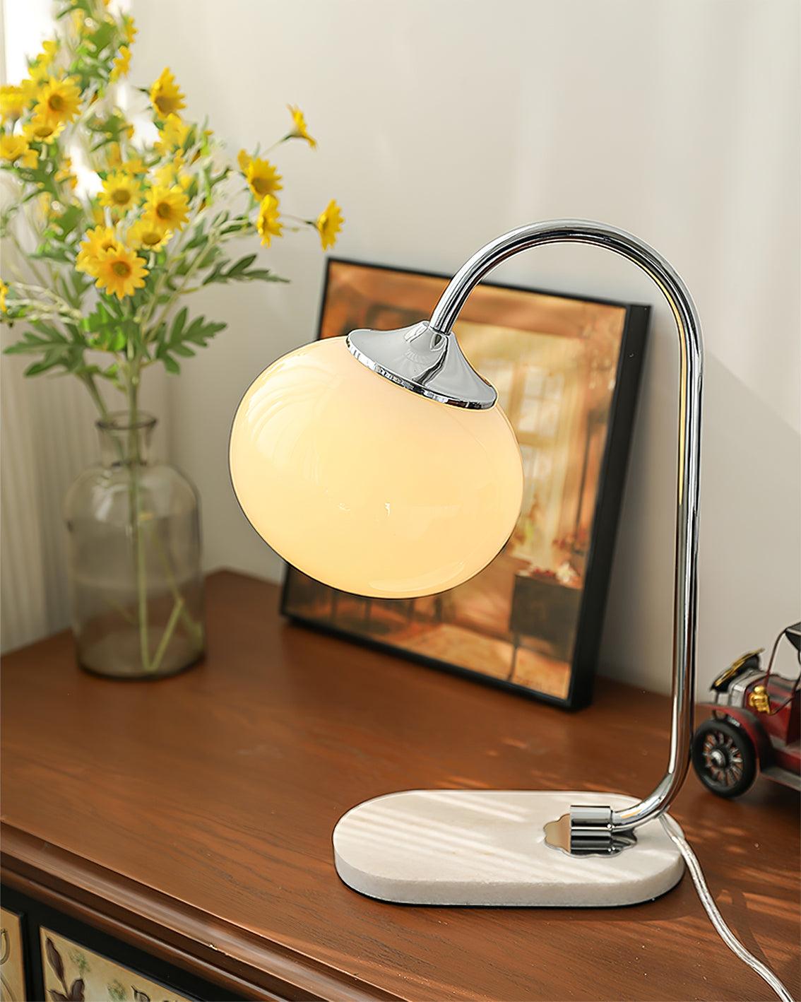 An off-white glass shade lights the Clérachic table lamp's chrome arm, set on a wooden dresser beside a framed picture and yellow daisies.