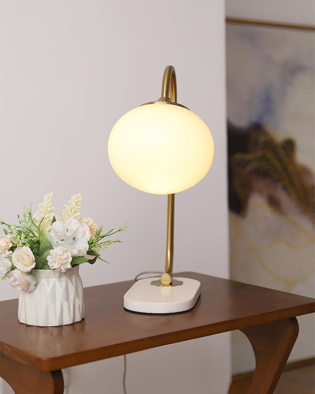 The Clérachic table lamp's white shade glows on a gold arm, placed on a wood side table beside a vase of small white flowers.