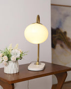 The Clérachic table lamp's white shade glows on a gold arm, placed on a wood side table beside a vase of small white flowers.