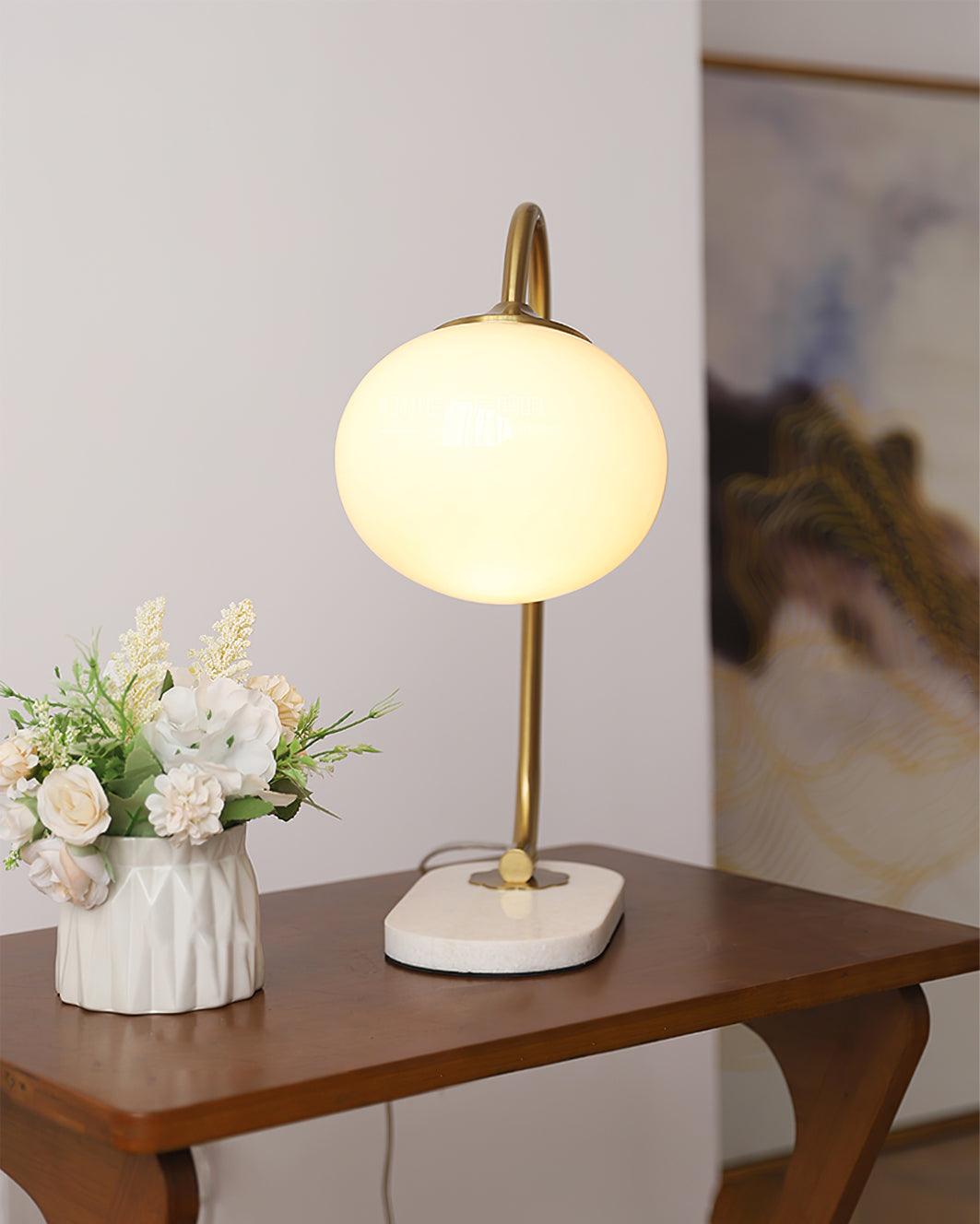 The Clérachic table lamp's white shade glows on a gold arm, placed on a wood side table beside a vase of small white flowers.