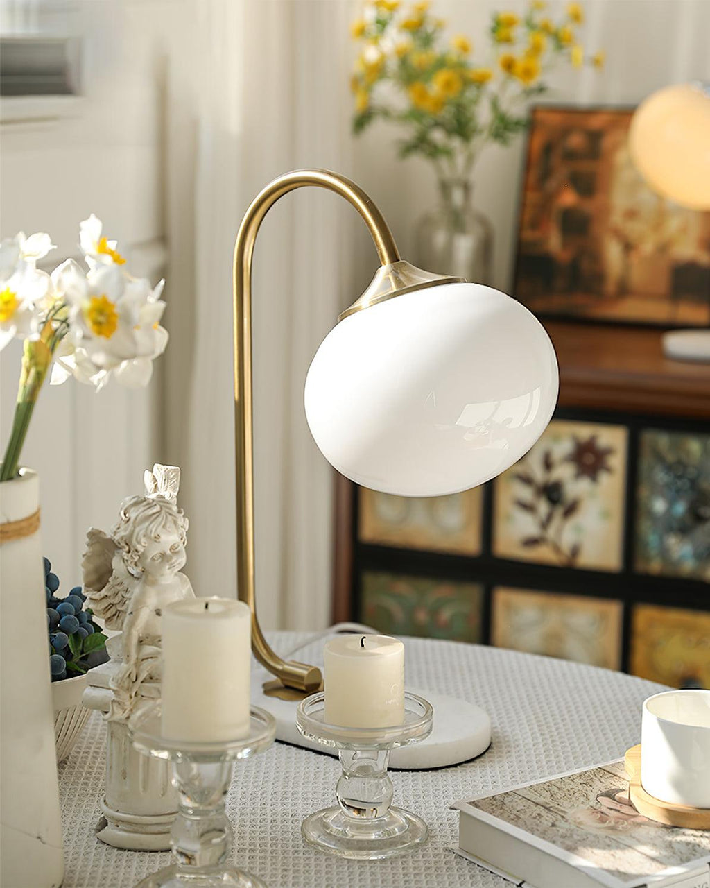 A lit Clérachic table lamp with white shade and gold arm sits among candles, stacked books and a small figurine on a dressed table.