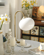 A lit Clérachic table lamp with white shade and gold arm sits among candles, stacked books and a small figurine on a dressed table.