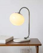 The Clérachic table lamp's white glass shade glows on a chrome arm, set on a wooden table beside a stack of books near a wall socket.