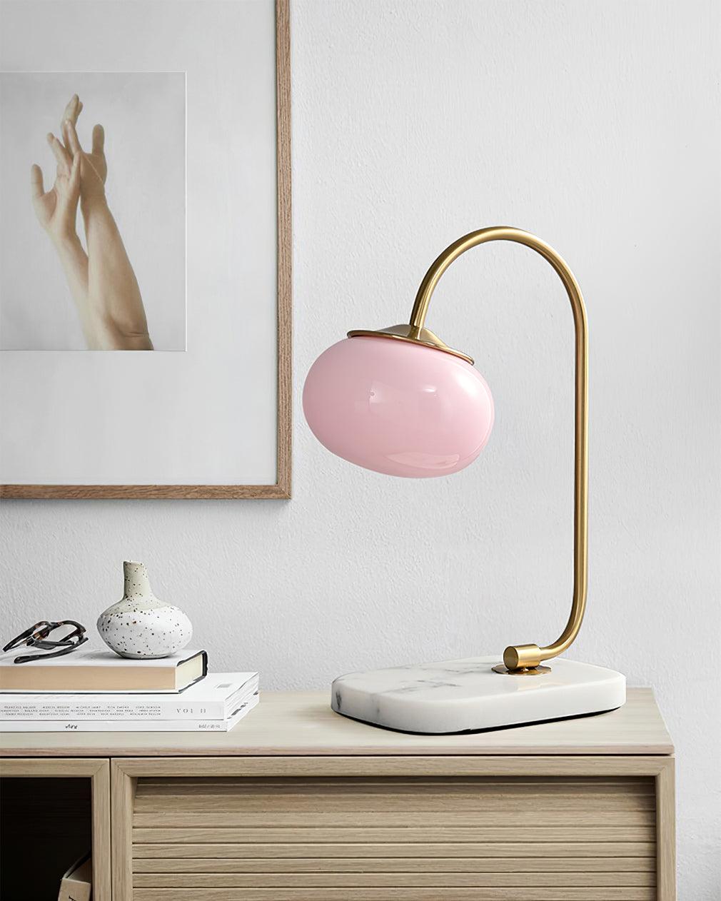 An unlit pink-shaded Clérachic table lamp on a gold arm stands on a wooden surface beside stacked books and a folded glasses case.