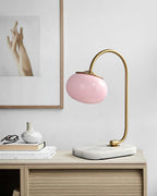 An unlit pink-shaded Clérachic table lamp on a gold arm stands on a wooden surface beside stacked books and a folded glasses case.