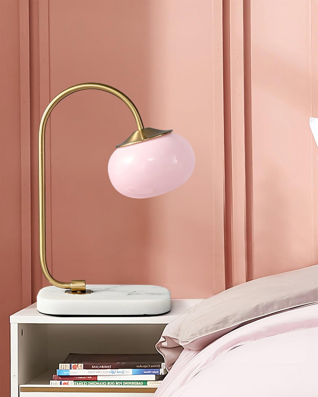The Clérachic table lamp's pink shade sits on a marble-topped nightstand against a dusty pink wall, folded bedding visible behind it.