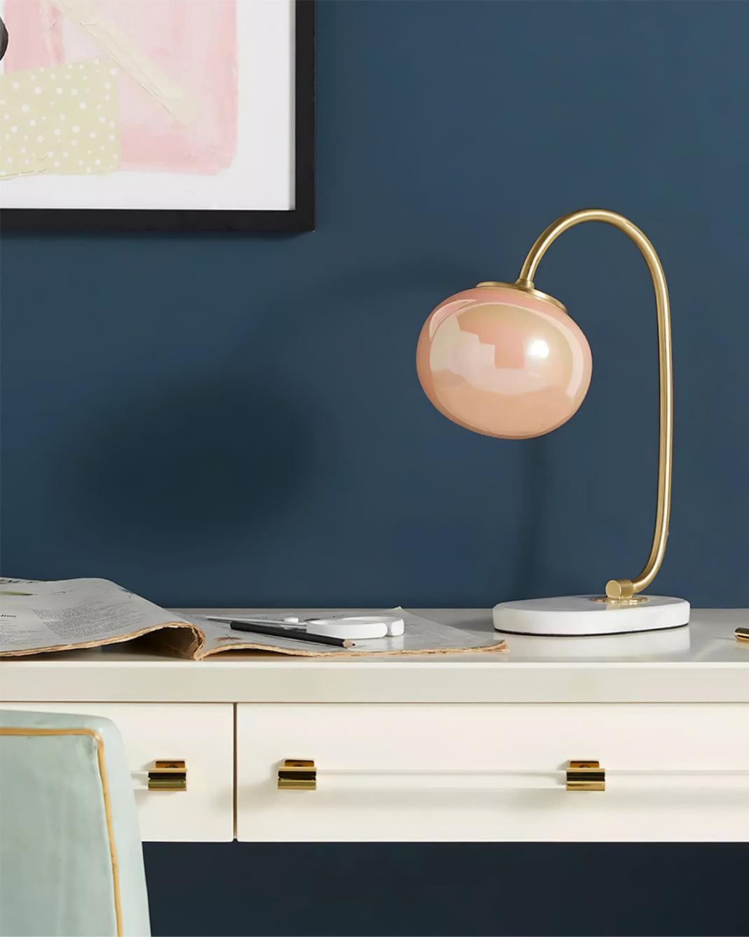 A peach glass shade tops the Clérachic table lamp's gold arm on a white dresser, papers and a pen resting against a navy wall.
