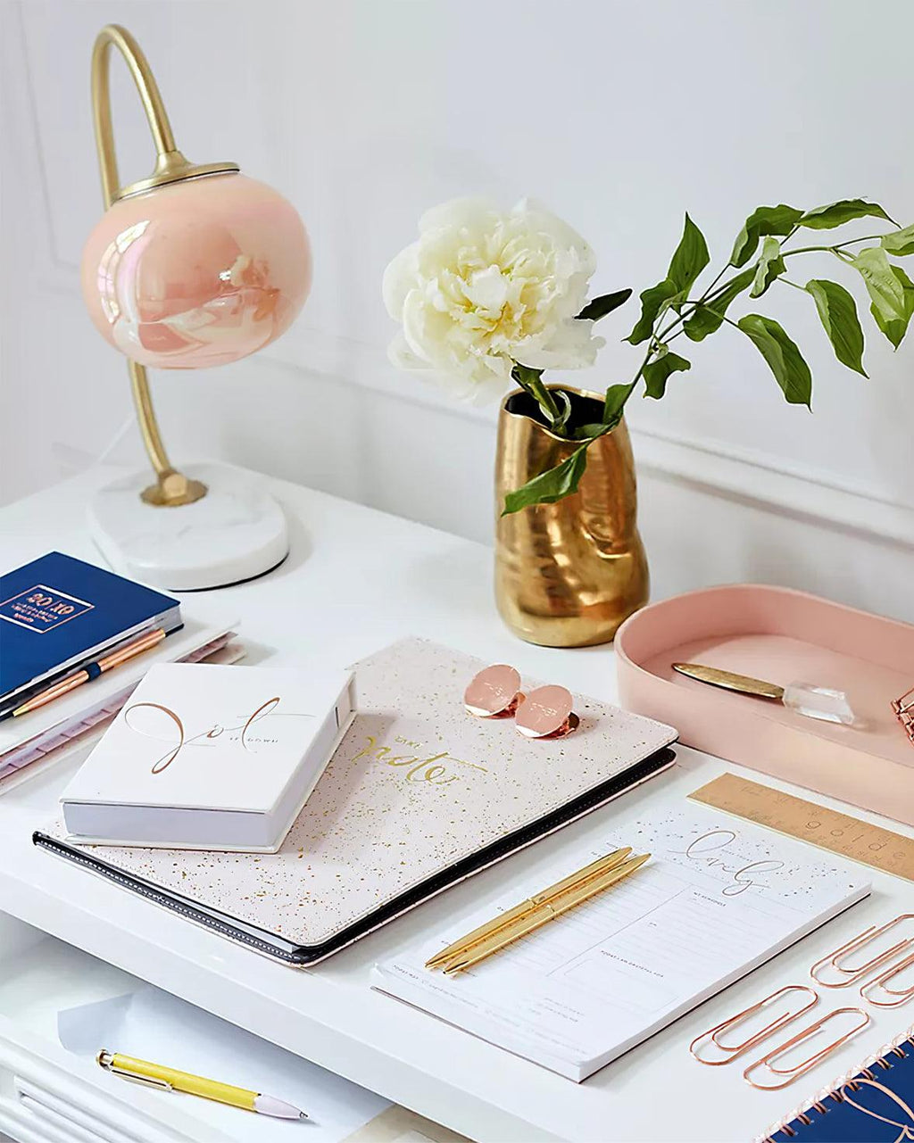 The Clérachic table lamp's marbled pink-peach shade glows on a white desk beside notebooks, a pink tray and a gold vase of flowers.