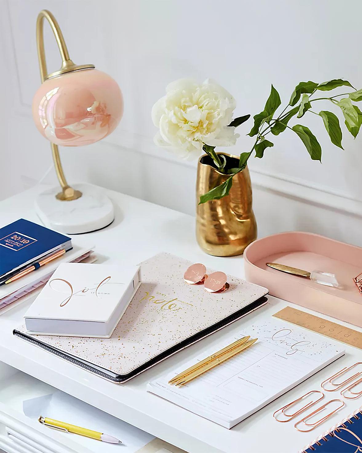 The Clérachic table lamp's marbled pink-peach shade glows on a white desk beside notebooks, a pink tray and a gold vase of flowers.