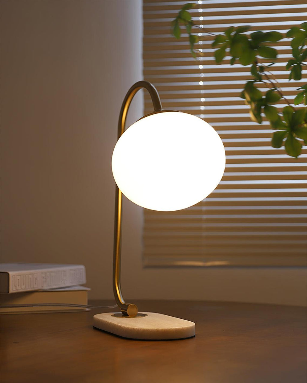 A lit Clérachic table lamp with brass arm and white shade stands on a round wooden base, morning light through blinds behind stacked books.