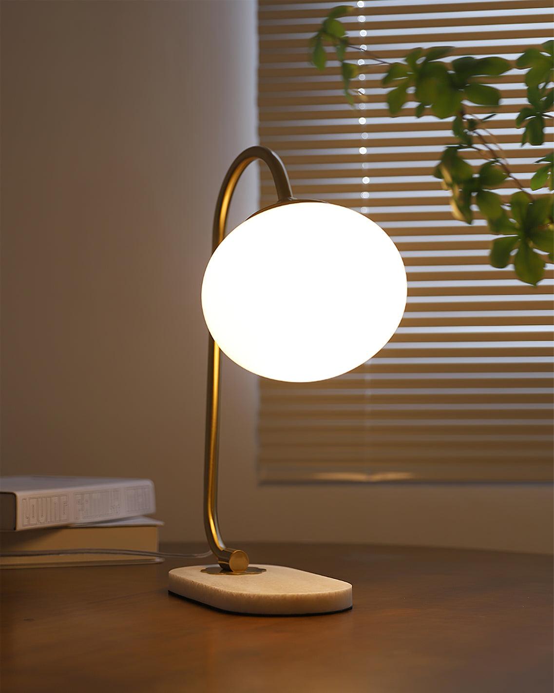 A lit Clérachic table lamp with brass arm and white shade stands on a round wooden base, morning light through blinds behind stacked books.