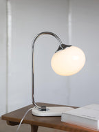 The Clérachic table lamp's white glass shade glows on a chrome arm, set on a rectangular marble base at a wooden table's edge beside a book.