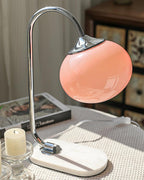 An unlit pink-shaded Clérachic table lamp on a chrome arm and marble base sits beside a candle, a patterned throw visible behind it.