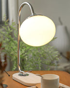 The Clérachic table lamp's white shade lights a chrome arm on a pale wooden board, greenery and a mug with a spoon nearby.