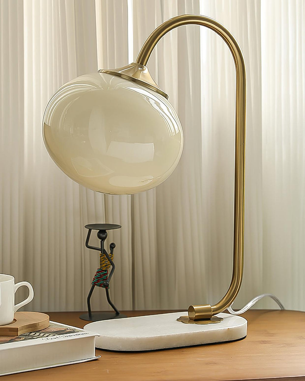 A champagne-toned glass shade tops the Clérachic table lamp's gold arm and marble base, beside a small figurine and a teacup on wood.
