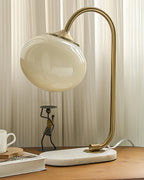 A champagne-toned glass shade tops the Clérachic table lamp's gold arm and marble base, beside a small figurine and a teacup on wood.
