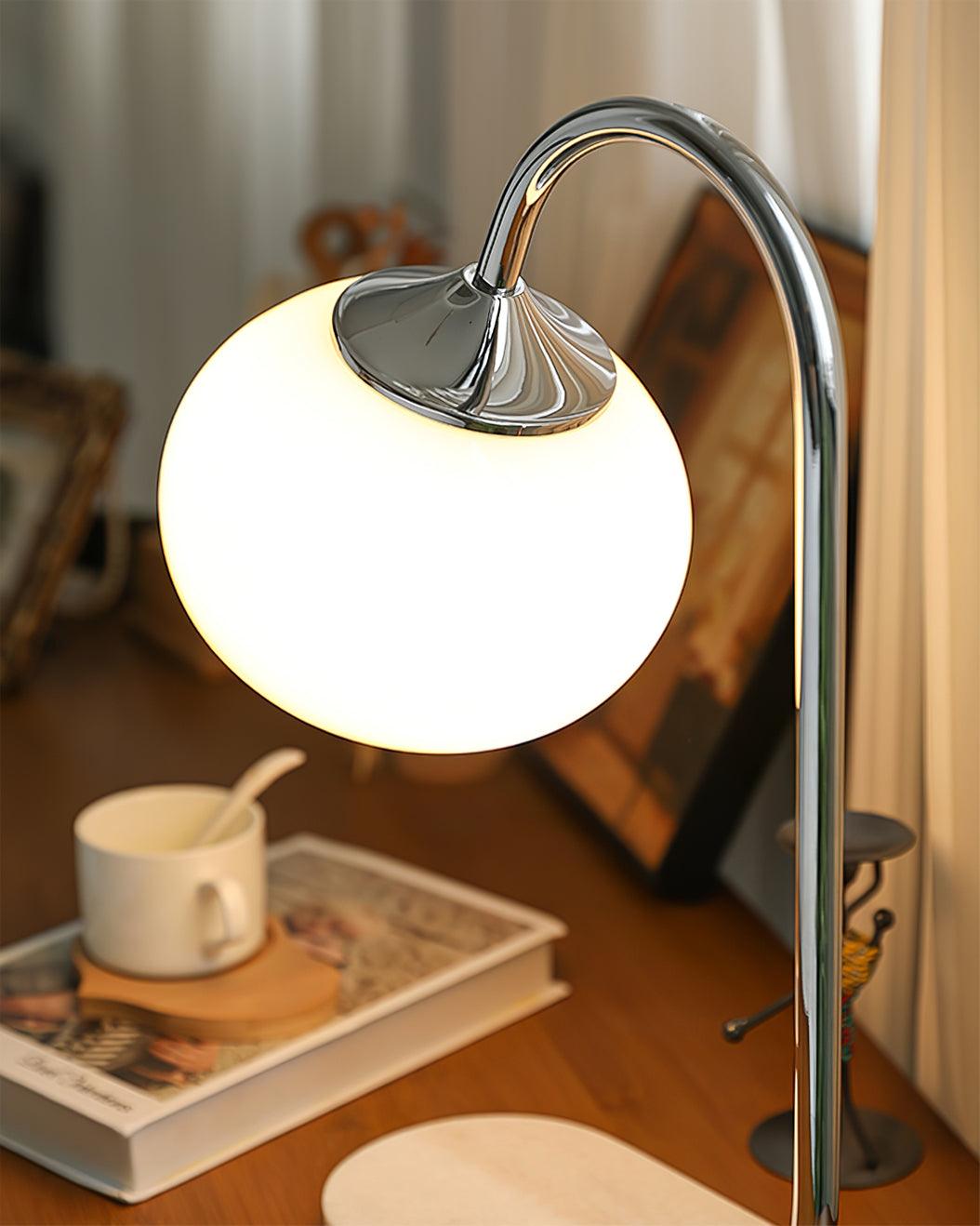 The Clérachic table lamp's white shade glows on a chrome arm and marble base, set on a wooden table beside a mug and stacked books.