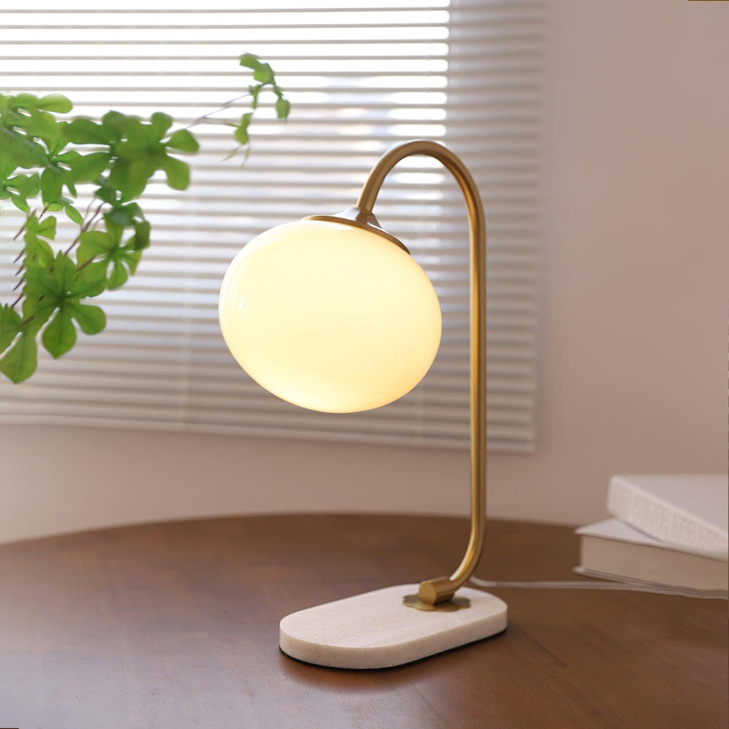 Sunlight through slatted blinds falls on a lit Clérachic table lamp, its brass arm and white shade standing on a wooden desk beside books.