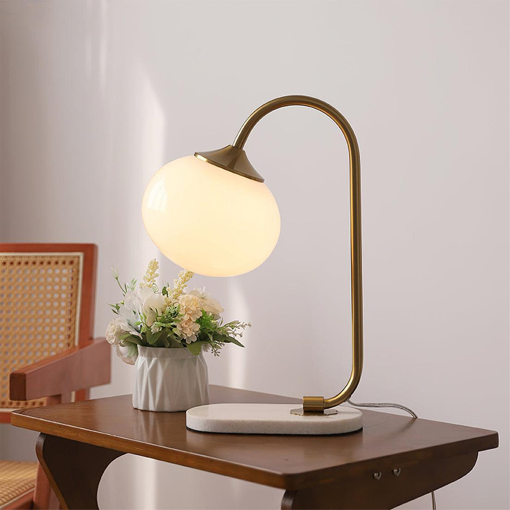The Clérachic table lamp's white glass shade glows warm on a brass arm, set on a cane-backed chair's side table beside a small potted plant.