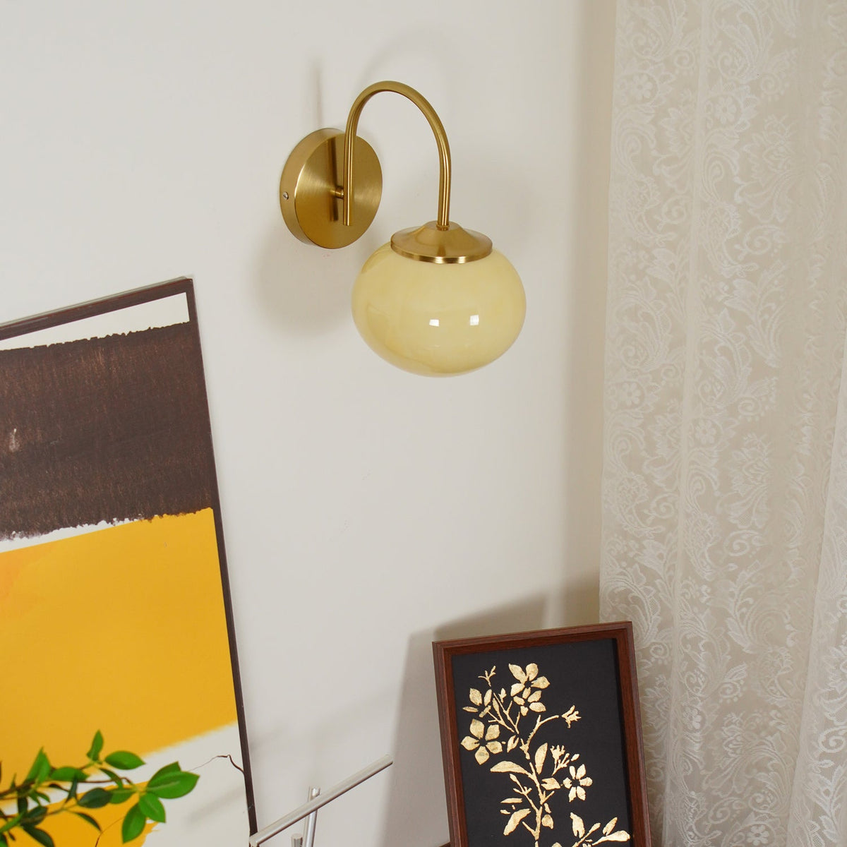 Framed floral art sits below this Clérachic wall light in copper and beige glass, mounted near sheer curtains.