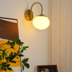 A framed picture and trailing greenery flank this Clérachic wall light in copper and white glass, lit, on a cream wall.