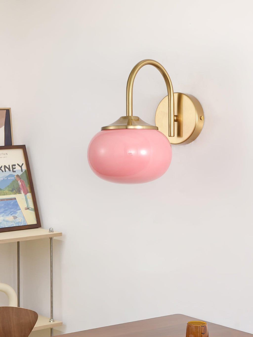 Framed art sits on a shelf beneath this Clérachic wall light in copper and pink glass, on a plain white wall.