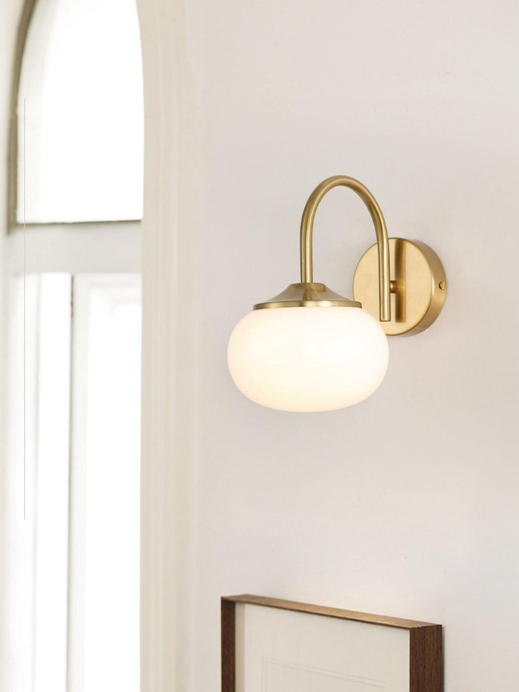 Near a window, this Clérachic wall light in copper and white glass hangs beside a framed picture.