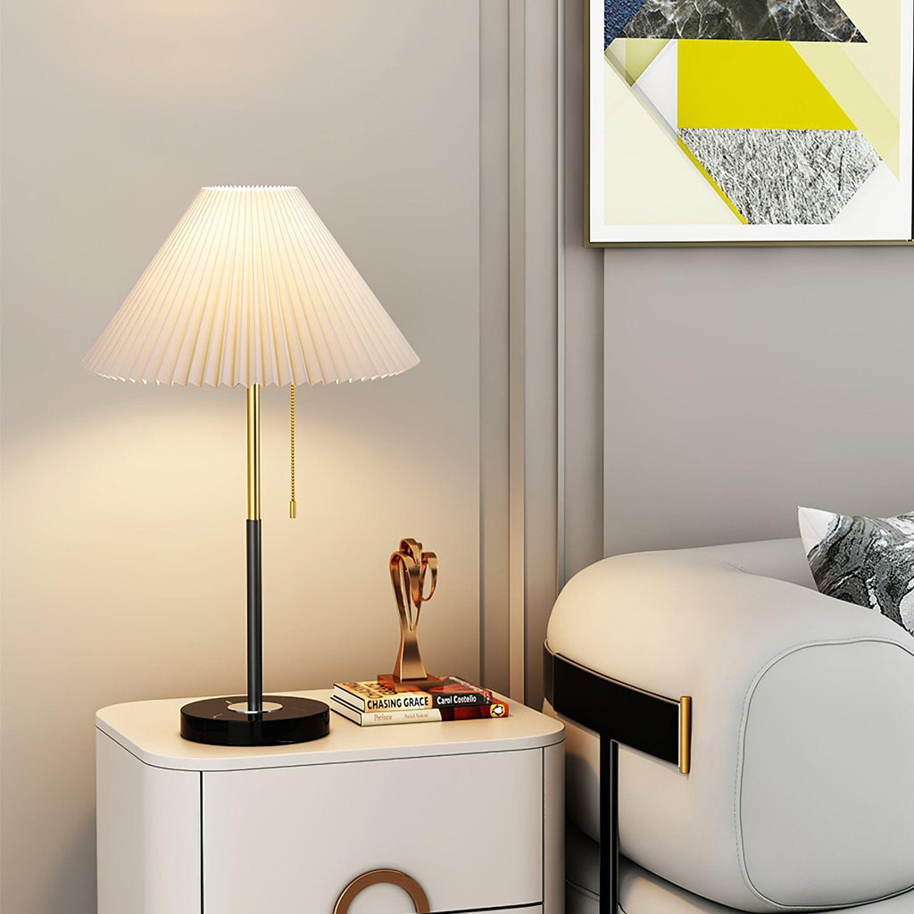 Clérachic lamp lit on a white bedside cabinet beside stacked books and a small sculpture, cream pleated shade glowing warm.