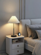 Clérachic lamp lit on a grey bedside cabinet with open shelving and drawers, an alarm clock beside it.
