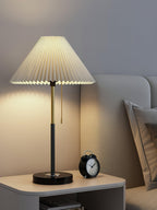 Clérachic lamp with its pull-cord switch, lit on a bedside cabinet with an alarm clock and pillows nearby.
