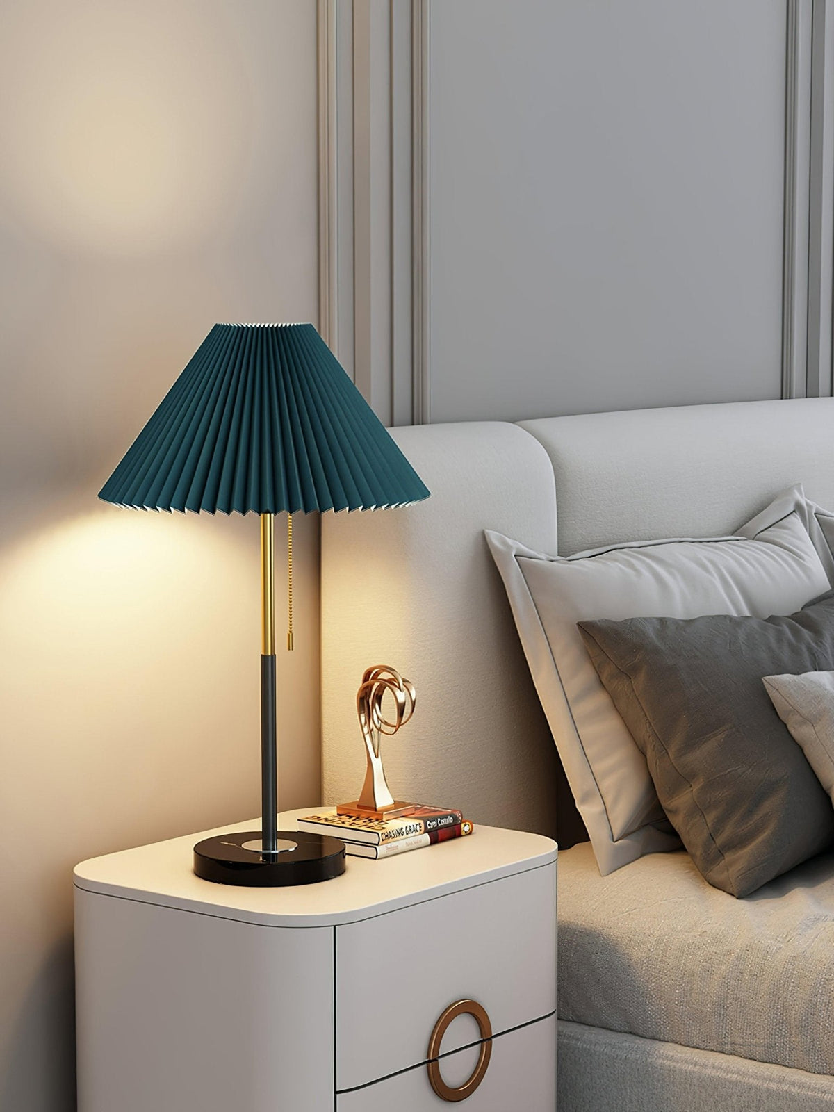 Clérachic lamp shown in the dark teal shade colourway, lit beside a small sculpture on the bedside cabinet.