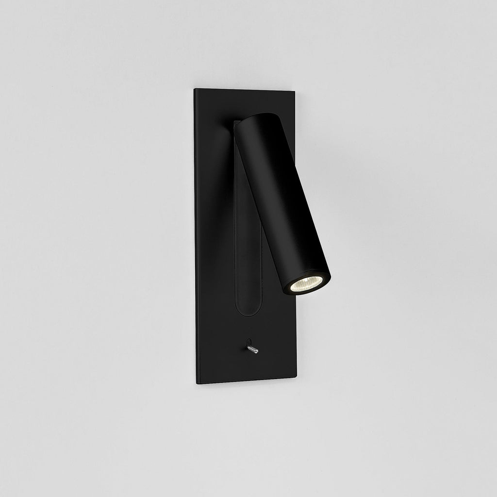 This Clérachic reading light in matt black, lit, mounted on a plain white wall, head angled down.