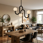 A black-finish chandelier with three glowing alabaster globes hangs above a wood dining table, styled with a round mirror and sofa nearby, from Clérachic.