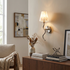 In a living room beside a console with books and a vase of dried branches, this Clérachic wall sconce in brushed nickel sits near a cream armchair.