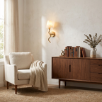 Beside a sunlit window, this Clérachic wall sconce in brushed brass warms a console with books and a vase near a cream armchair.
