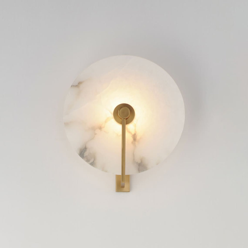 Close view of this Clérachic wall sconce in brushed brass, its round alabaster disc lit warm on a grey wall.